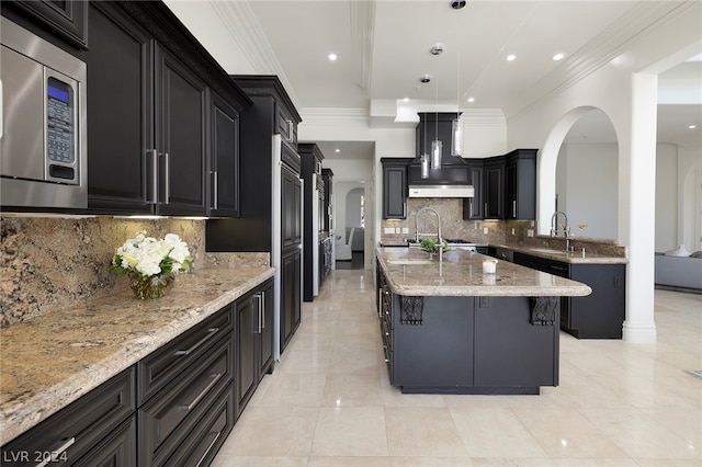 kitchen featuring stainless steel microwave, decorative light fixtures, ornamental molding, a kitchen island with sink, and light stone countertops