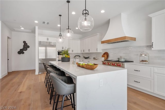 kitchen featuring white cabinetry, stainless steel appliances, premium range hood, hanging light fixtures, and a center island with sink