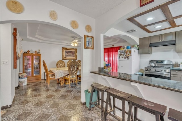 kitchen featuring ceiling fan, stainless steel gas stove, coffered ceiling, white refrigerator, and dark stone countertops