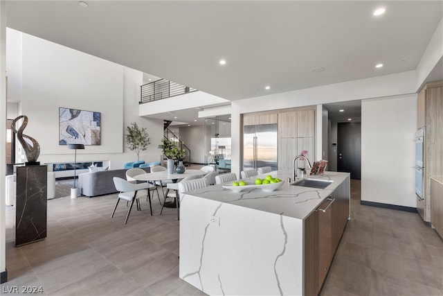 kitchen with light stone countertops, sink, double oven, an island with sink, and stainless steel built in fridge