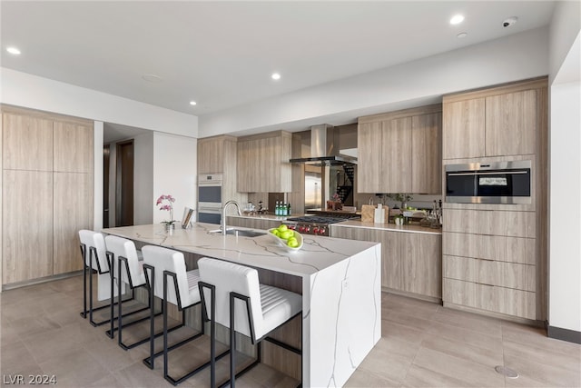 kitchen featuring a kitchen island with sink, wall chimney range hood, sink, appliances with stainless steel finishes, and light stone counters