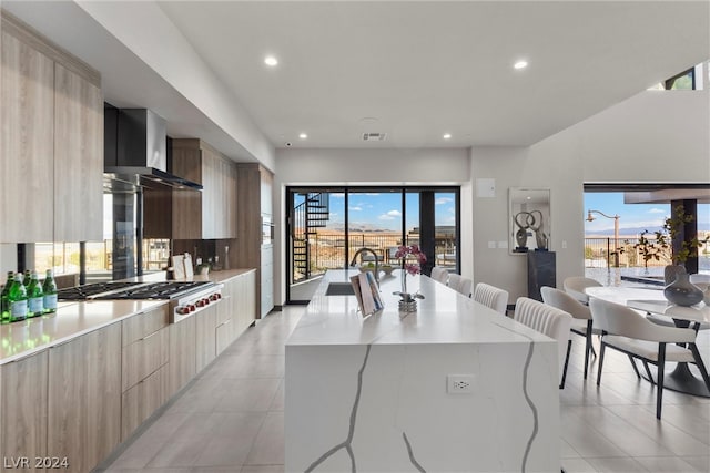 kitchen featuring light stone countertops, a kitchen island with sink, wall chimney range hood, light tile patterned floors, and stainless steel gas stovetop