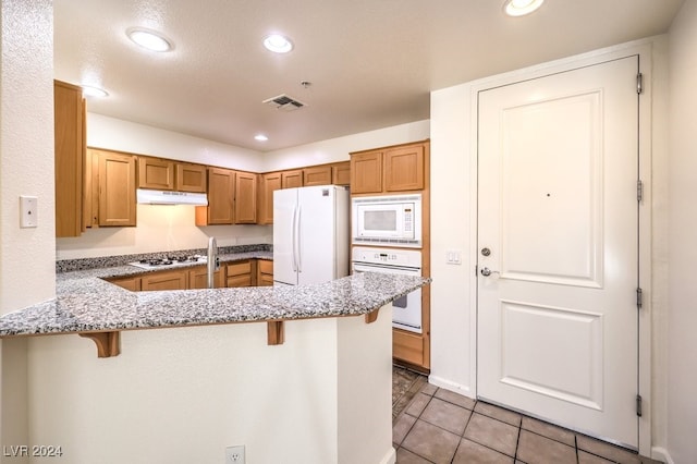 kitchen featuring kitchen peninsula, white appliances, a kitchen bar, and light tile patterned flooring