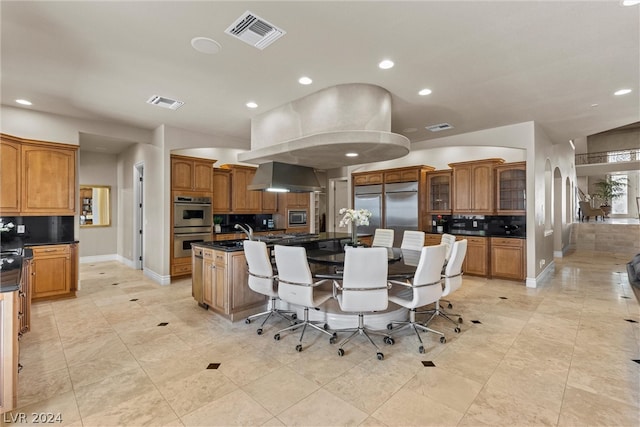 kitchen with wall chimney range hood, appliances with stainless steel finishes, light tile patterned floors, backsplash, and a kitchen island with sink