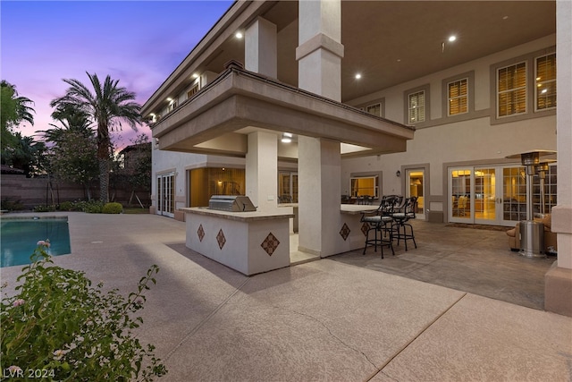 patio terrace at dusk with a balcony, exterior kitchen, an outdoor bar, and a fenced in pool