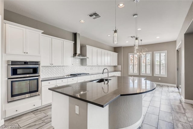 kitchen featuring wall chimney range hood, pendant lighting, a center island with sink, sink, and white cabinets