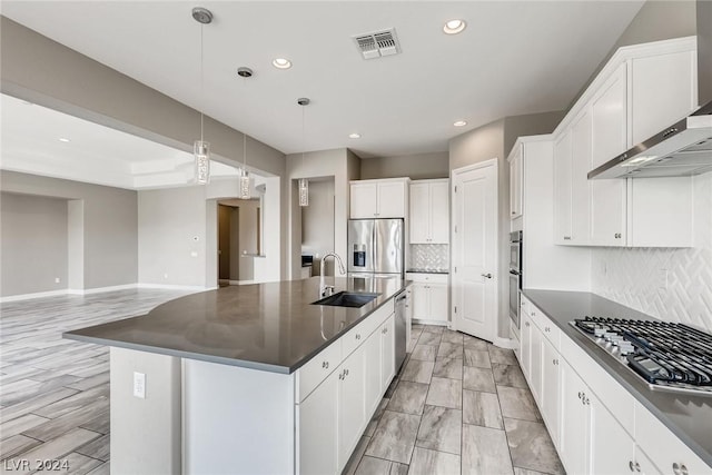 kitchen featuring decorative light fixtures, wall chimney range hood, a center island with sink, sink, and appliances with stainless steel finishes