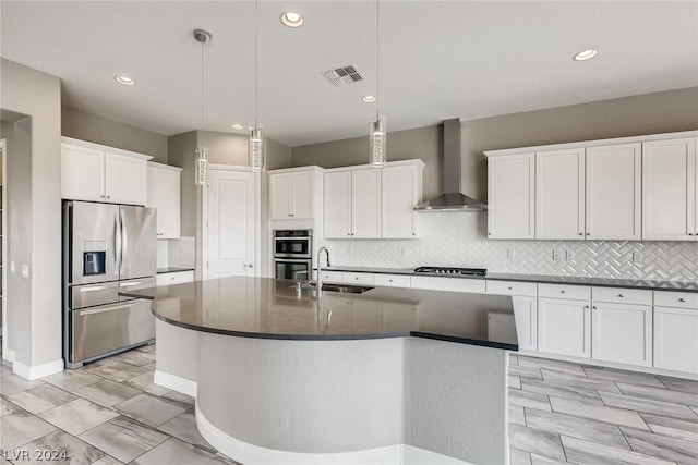 kitchen featuring white cabinets, wall chimney exhaust hood, stainless steel appliances, hanging light fixtures, and a center island with sink