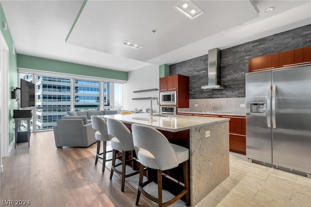 kitchen with wall chimney range hood, light hardwood / wood-style flooring, an island with sink, stainless steel appliances, and a breakfast bar area