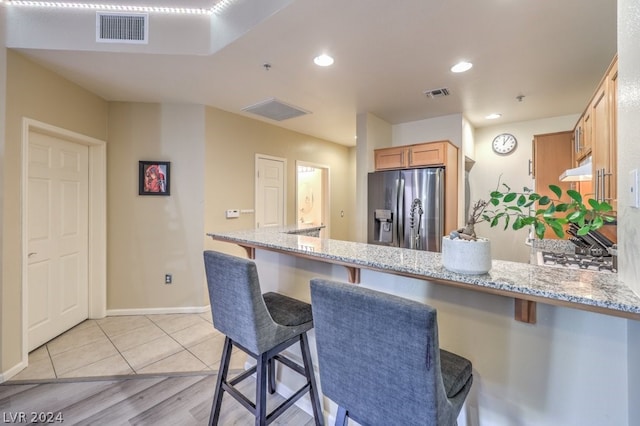 kitchen featuring light stone countertops, kitchen peninsula, stainless steel fridge, light tile floors, and a kitchen bar