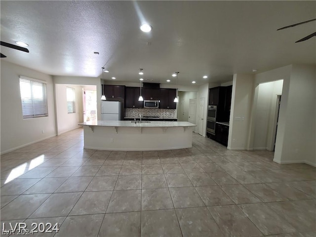 kitchen featuring ceiling fan, light tile patterned floors, an island with sink, appliances with stainless steel finishes, and tasteful backsplash