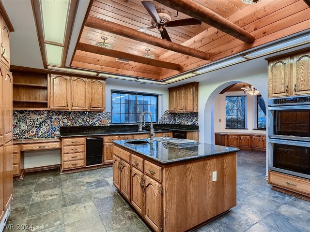 kitchen featuring ceiling fan, backsplash, stainless steel double oven, a kitchen island with sink, and dark tile floors