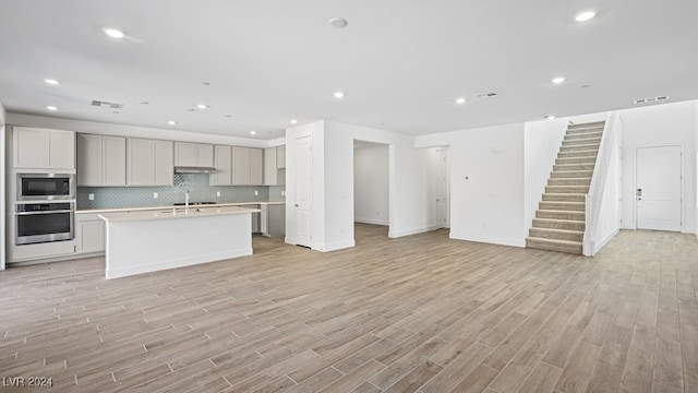 kitchen featuring built in microwave, an island with sink, oven, light hardwood / wood-style floors, and backsplash