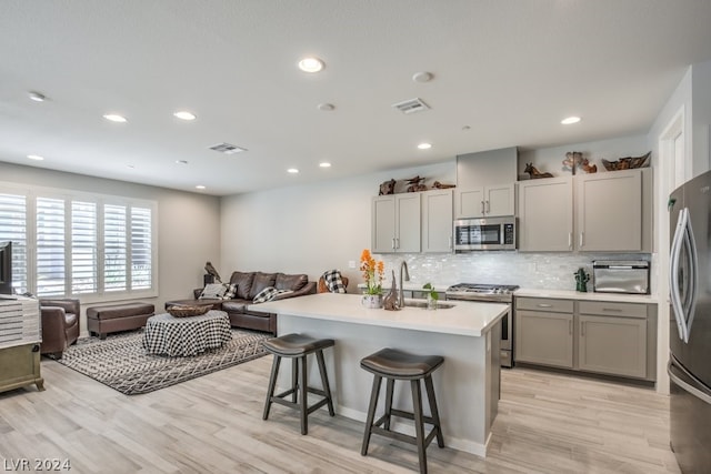 kitchen with a breakfast bar, backsplash, a center island with sink, light wood-type flooring, and appliances with stainless steel finishes