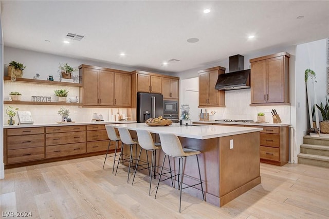 kitchen with wall chimney exhaust hood, light wood-type flooring, a breakfast bar, a center island with sink, and stainless steel appliances