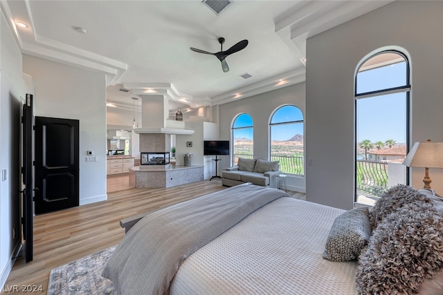 bedroom with light wood-type flooring, access to outside, a tray ceiling, a tile fireplace, and ceiling fan