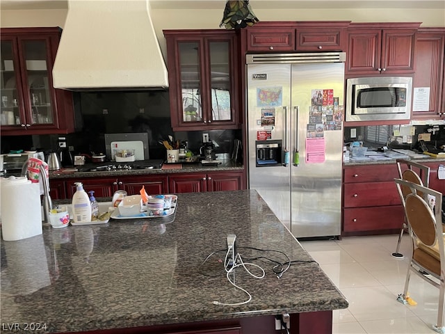 kitchen featuring tasteful backsplash, custom exhaust hood, built in appliances, dark stone countertops, and light tile floors