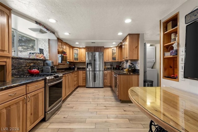 kitchen with light wood-type flooring, stacked washer / drying machine, dark stone countertops, a textured ceiling, and appliances with stainless steel finishes