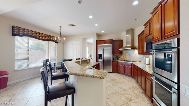 kitchen with sink, built in appliances, pendant lighting, a kitchen island with sink, and wall chimney range hood