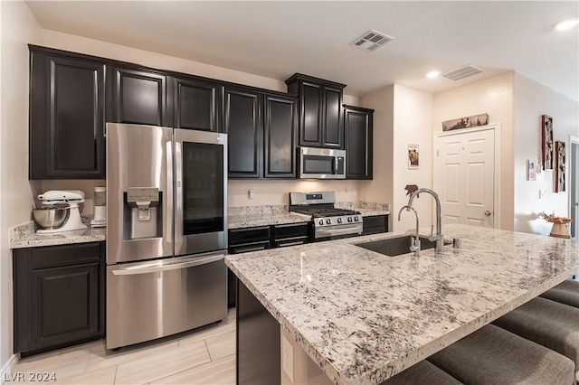 kitchen featuring a kitchen island with sink, sink, light stone countertops, appliances with stainless steel finishes, and a kitchen bar