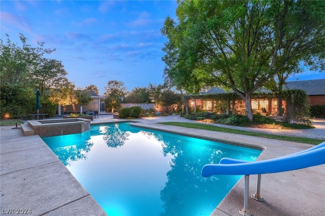 pool at dusk with a patio area, a water slide, and an in ground hot tub