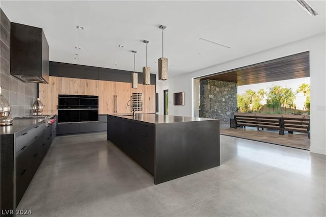 kitchen featuring wall chimney exhaust hood, hanging light fixtures, black double oven, an island with sink, and stainless steel gas stovetop