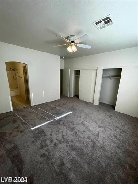 unfurnished bedroom with ceiling fan, ensuite bath, multiple closets, and dark colored carpet