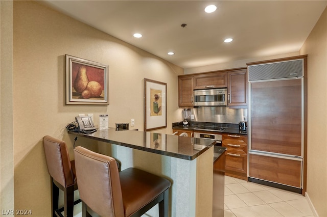 kitchen featuring kitchen peninsula, light tile patterned floors, a kitchen bar, dark stone countertops, and stainless steel appliances