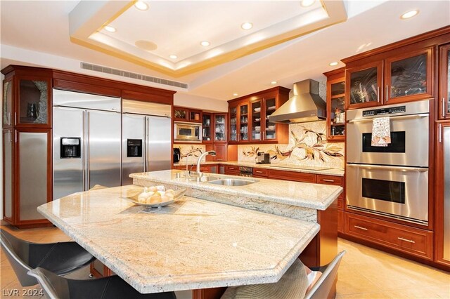 kitchen with built in appliances, a raised ceiling, wall chimney exhaust hood, and an island with sink