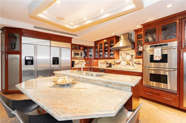 kitchen with a raised ceiling, wall chimney exhaust hood, a breakfast bar area, built in appliances, and a sink