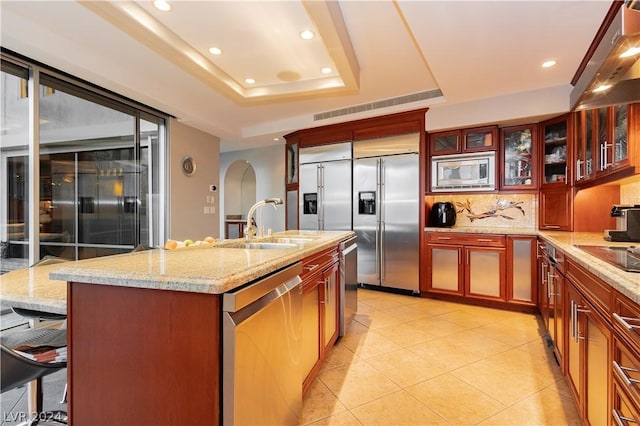 kitchen with a tray ceiling, tasteful backsplash, an island with sink, light stone countertops, and built in appliances