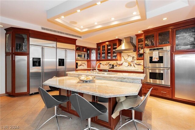 kitchen featuring built in appliances, a raised ceiling, wall chimney exhaust hood, and a kitchen island with sink