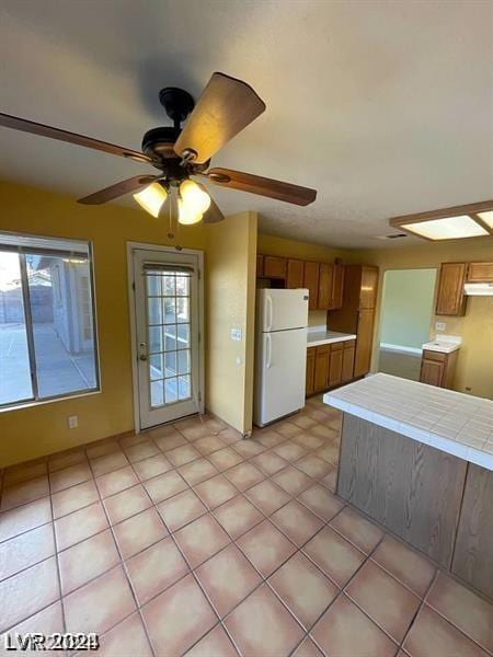 kitchen featuring a ceiling fan, under cabinet range hood, freestanding refrigerator, tile countertops, and brown cabinetry