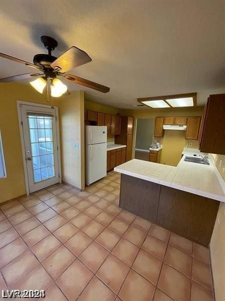 kitchen with a ceiling fan, brown cabinetry, a peninsula, freestanding refrigerator, and under cabinet range hood