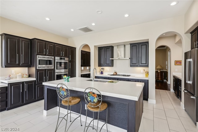 kitchen with sink, a breakfast bar, appliances with stainless steel finishes, a kitchen island with sink, and wall chimney range hood