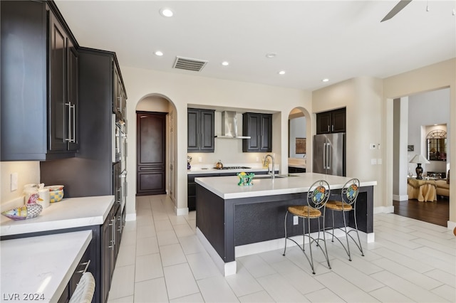 kitchen with wall chimney range hood, appliances with stainless steel finishes, a breakfast bar area, ceiling fan, and a kitchen island with sink