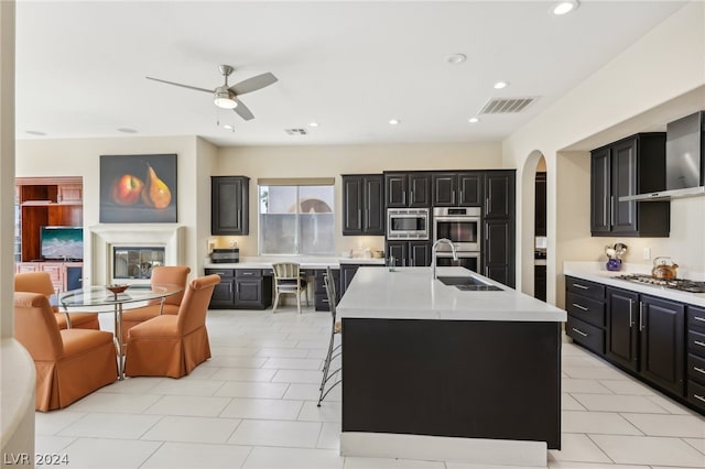 kitchen with a center island with sink, wall chimney range hood, appliances with stainless steel finishes, sink, and ceiling fan