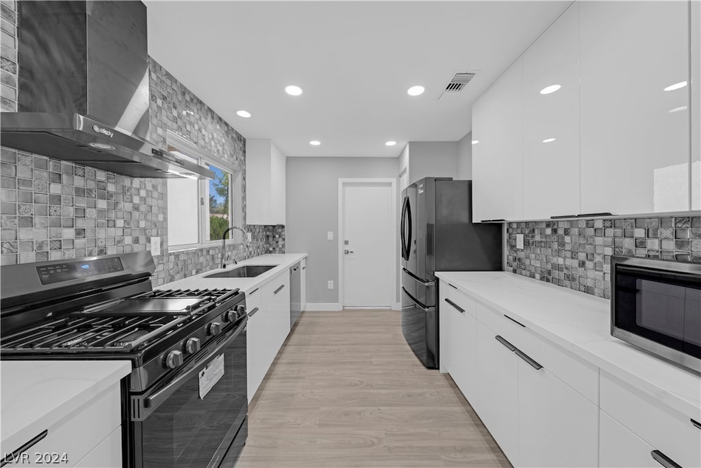 kitchen with sink, wall chimney range hood, white cabinetry, appliances with stainless steel finishes, and light hardwood / wood-style floors