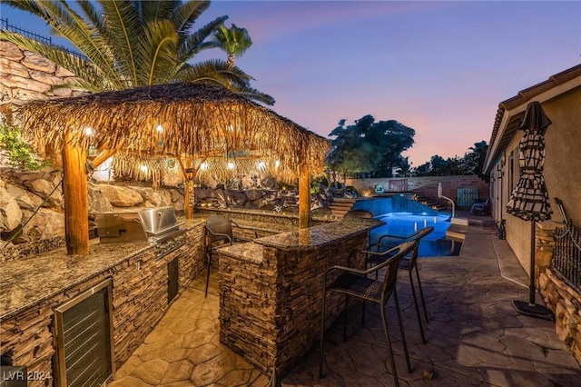 patio terrace at dusk with a fenced in pool, area for grilling, exterior kitchen, and an outdoor bar