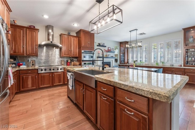 kitchen with decorative light fixtures, backsplash, wall chimney range hood, stainless steel appliances, and a center island with sink