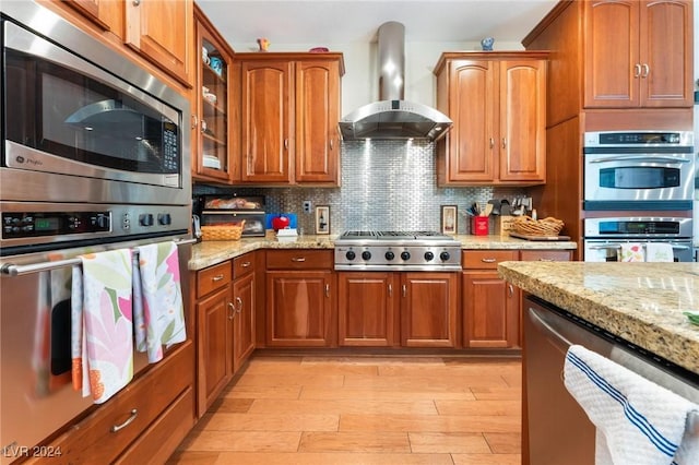 kitchen with tasteful backsplash, stainless steel appliances, light stone countertops, wall chimney range hood, and light hardwood / wood-style flooring