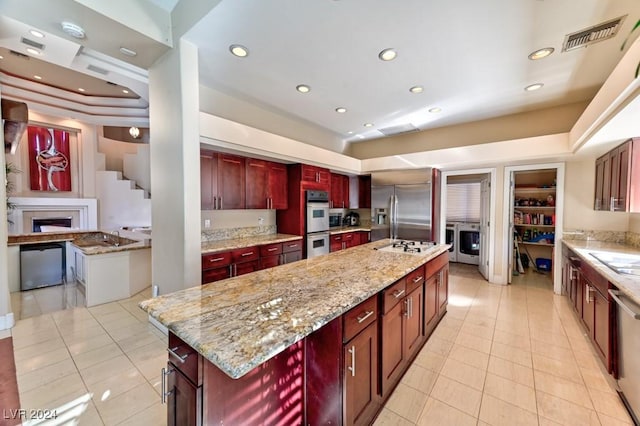 kitchen featuring appliances with stainless steel finishes, a tray ceiling, light stone countertops, a center island with sink, and washer / dryer