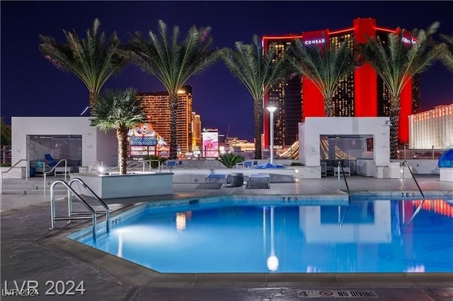 pool at twilight with a view of city lights, a patio, and a community pool