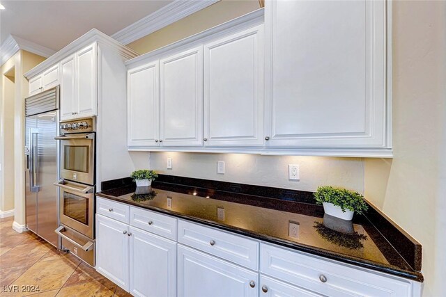 kitchen with appliances with stainless steel finishes, light tile patterned floors, dark stone countertops, white cabinetry, and ornamental molding