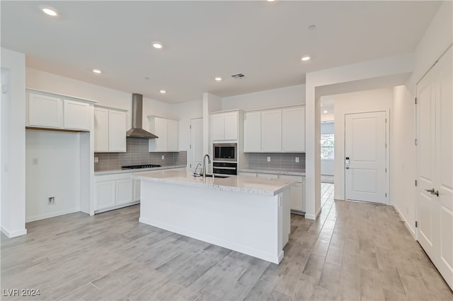 kitchen featuring a kitchen island with sink, appliances with stainless steel finishes, white cabinets, wall chimney exhaust hood, and light hardwood / wood-style flooring