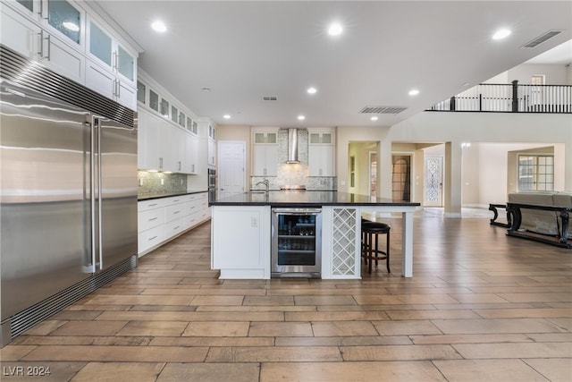 kitchen with built in refrigerator, beverage cooler, white cabinets, and wall chimney range hood