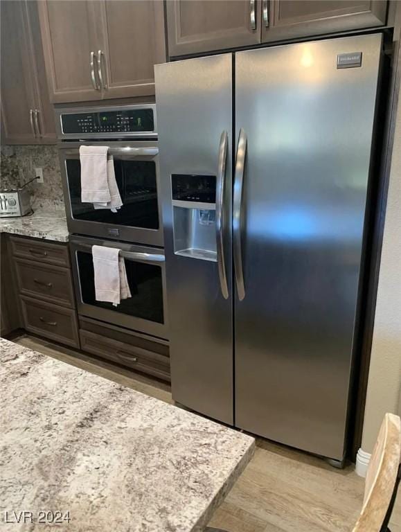 kitchen with tasteful backsplash, dark brown cabinets, appliances with stainless steel finishes, light stone countertops, and light hardwood / wood-style floors