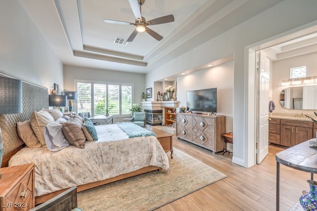 bedroom with visible vents, a glass covered fireplace, light wood-style flooring, a tray ceiling, and a sink