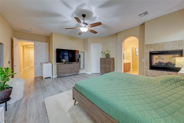 bedroom with a tiled fireplace, ceiling fan, ensuite bath, and light hardwood / wood-style floors