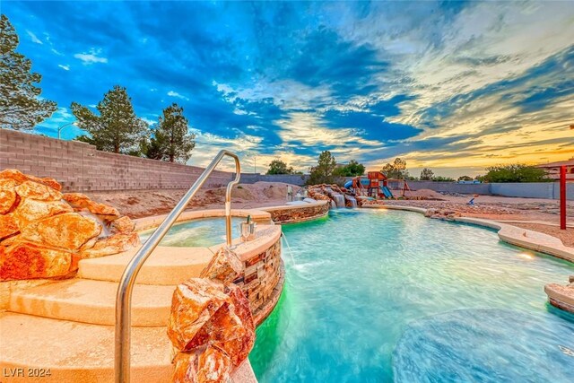 pool at dusk featuring a playground, pool water feature, and an in ground hot tub